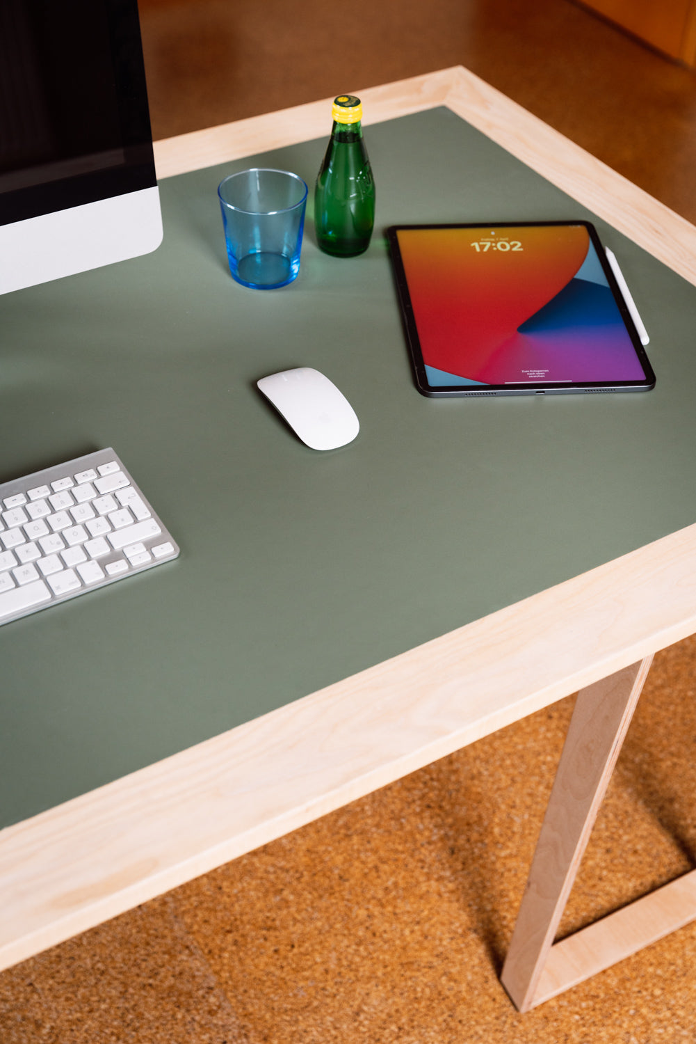 Minimalistischer Arbeitsplatz mit iMac-Monitor, weißer Tastatur, Magic Mouse, iPad mit Stift, grüne Glasflasche und blauem Becher auf einem hellen Holztisch im Büro. Minalistisches Design, hochwertige Materialien und ökologische Oberfläche