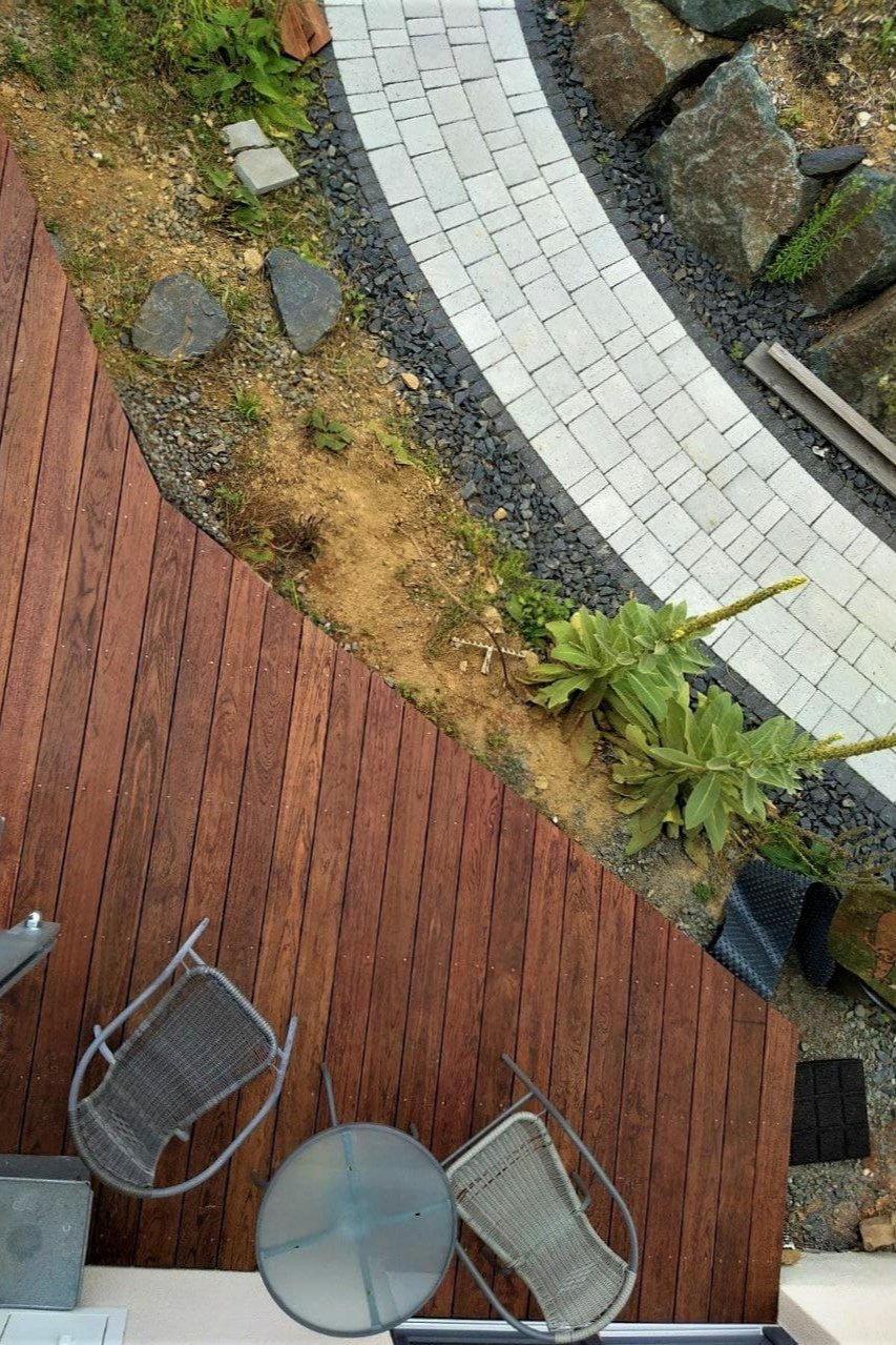 Vogelperspektive auf eine Terrasse mit Holzboden, zwei Metall-Stühlen, Glastisch, Pflanzenstreifen und geschwungenem Kiesweg, passend zu Holzfabrikats nachhaltigem Möbeldesign. Mediterane Stimmung, schönes Design, architektonisch