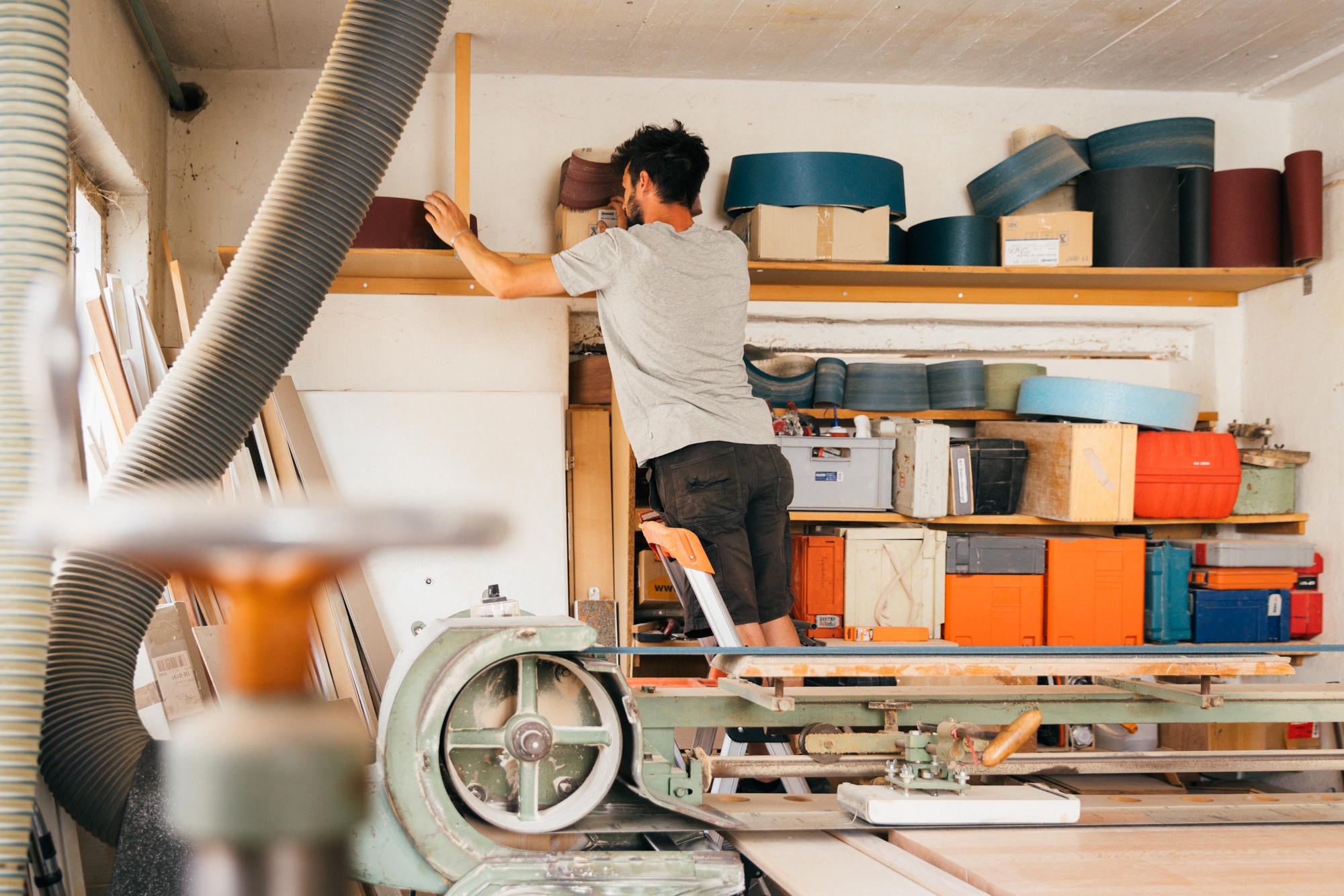 Ein Schreiner wählt Schleifpapier aus, während eine große Schleifmaschine in einer hellen, tageslichtdurchfluteten Werkstatt zu sehen ist.