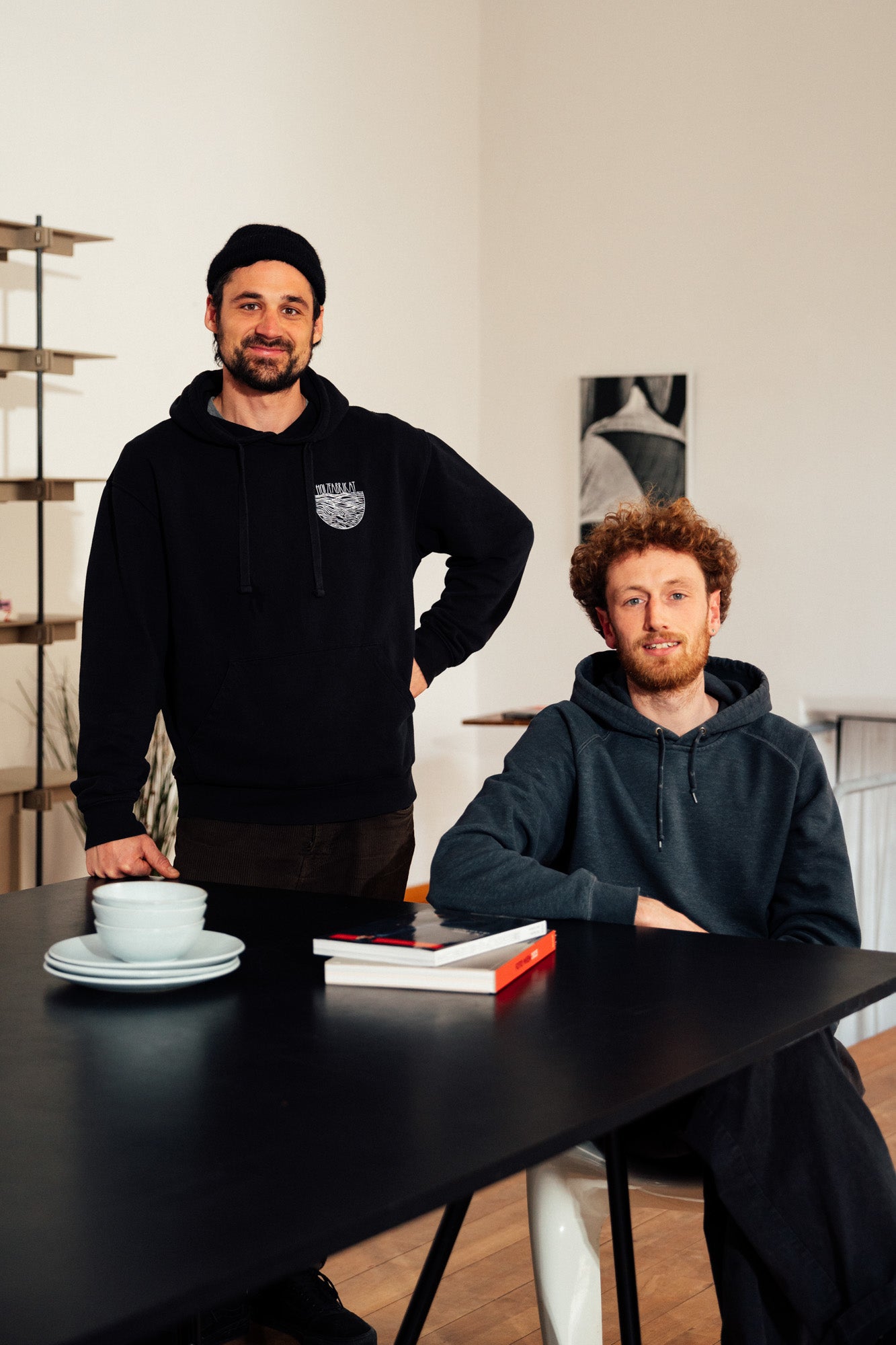 Zwei Männer, beide in dunklen Kapuzenpullovern, stehen und sitzen an einem schwarzen Tisch in einem hellen Raum. Auf dem Tisch liegen zwei Bücher sowie gestapelte Schüsseln und Teller. Die Männer sind Schreiner und Designer der Firma Holzfabrikat. Im Hintergrund ist das von ihnen entworfene Regalsystem „Tight“ zu sehen.