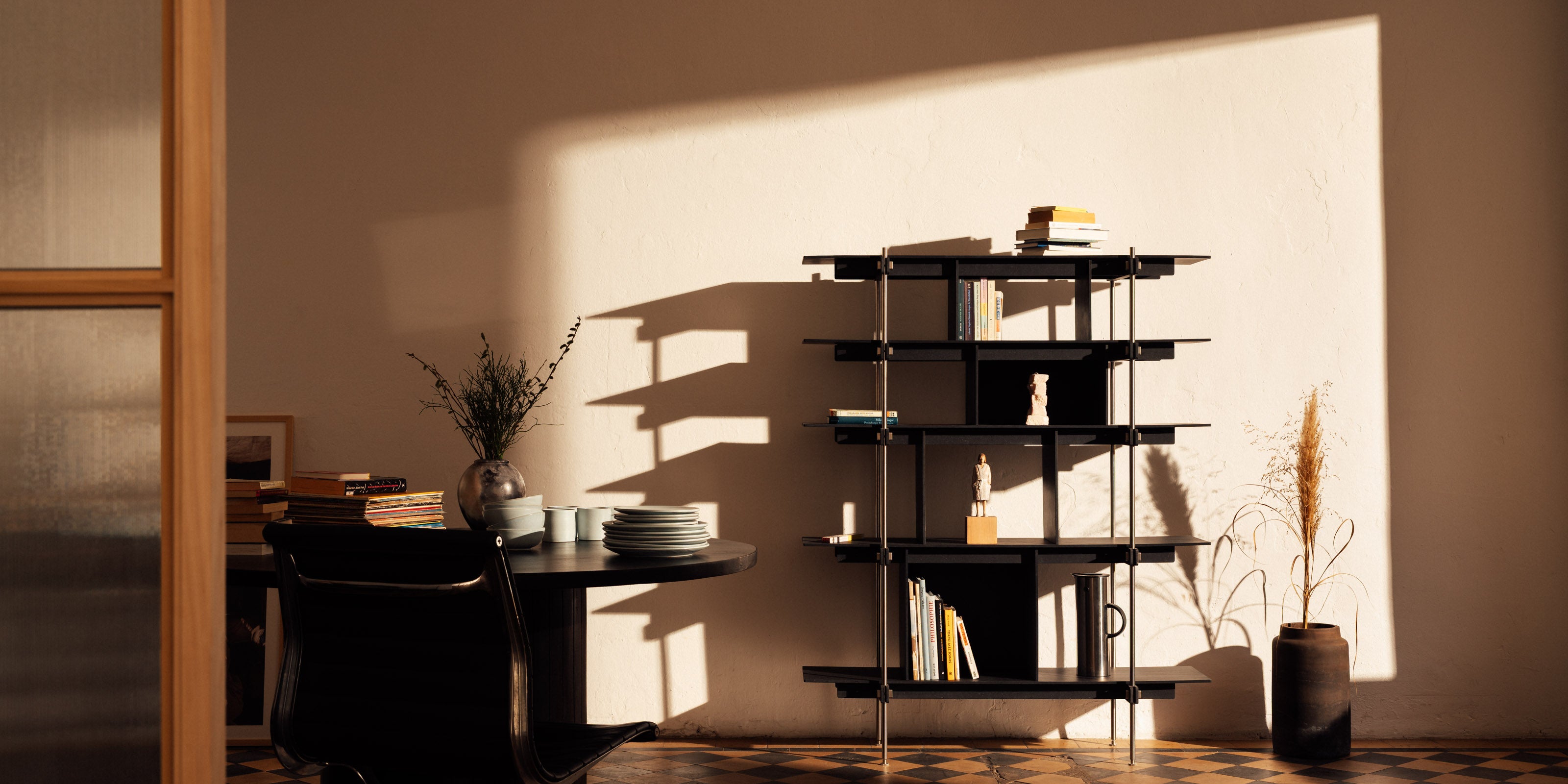 Ein modernes Design-Regalsystem aus schwarzem, durchgefärbtem MDF holz. Das Regal ist stilvoll mit Büchern und einer markanten Holzskulptur dekoriert und steht auf einem auffälligen Schachbrettboden.